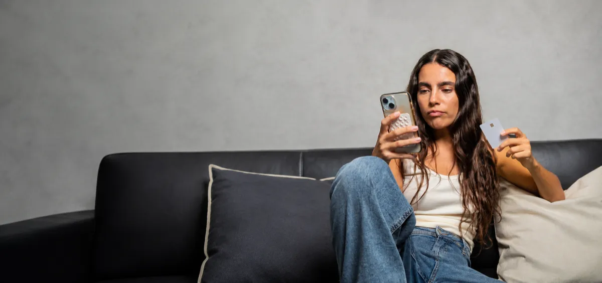 A woman sitting on a sofa looking at her phone, a payment card in her other hand and a pack of tablets held against the phone